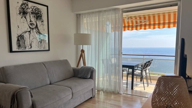 A modern living room with a grey sofa, a lamp, and a view of the sea through a balcony. - Incredible ocean view, pool, HVAC, Wi-Fi, sleeps 4 (Vallauris)
