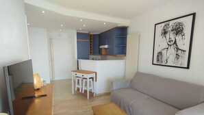 A modern living room with a sofa, a wooden table, and a TV. There is a kitchenette in the background with blue cabinets and a small island. - Incredible ocean view, pool, HVAC, Wi-Fi, sleeps 4 (Vallauris)