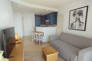 Ein modernes Wohnzimmer mit einer Couch, einem Holztisch und einem Fernseher. Im Hintergrund ist eine Küchenzeile mit blauen Schränken und einer kleinen Kochinsel zu sehen.