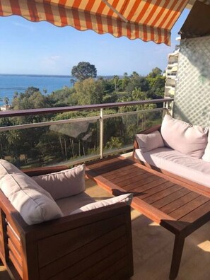 A balcony with a view of the ocean, a wooden table, and a sofa with cushions.