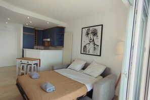 A hotel room with a bed, a desk, a chair, and a kitchenette in the background.