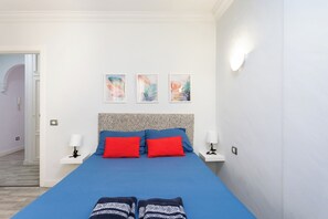 Ein Schlafzimmer mit einem blauen Bett, zwei roten Kissen, einem gemusterten Kopfteil und zwei Nachttischen mit Lampen. An der Wand hängen drei gerahmte Kunstwerke.