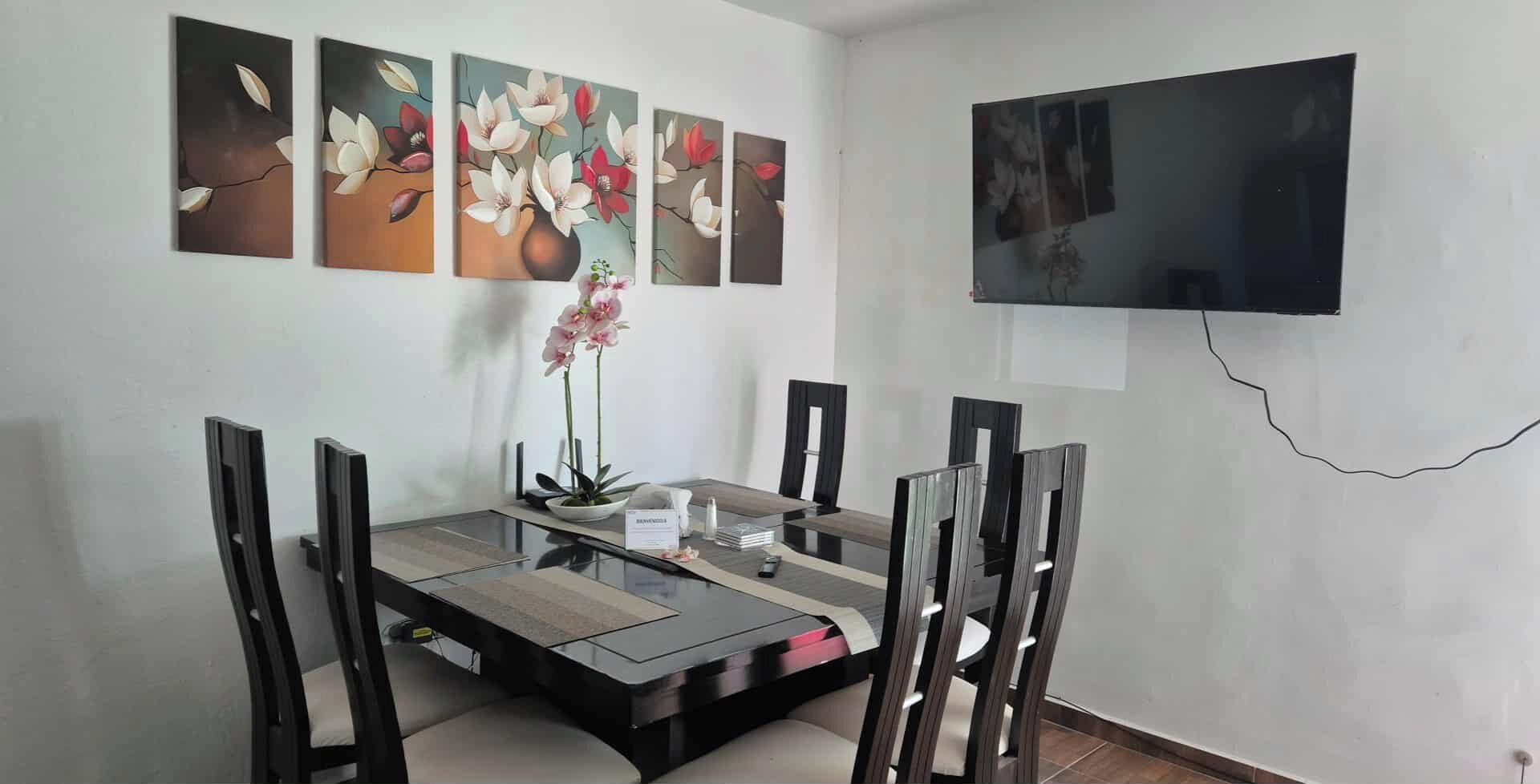 A dining area with a glass-top table, four chairs, a vase with flowers, and a flat-screen TV mounted on the wall.