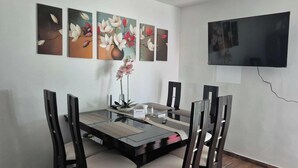 A dining area with a glass-top table, four chairs, a vase with flowers, and a flat-screen TV mounted on the wall.