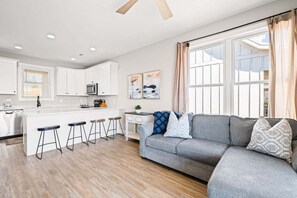 Eine moderne Küche mit weißen Schränken, einer zentralen Kochinsel mit Sitzgelegenheiten und einem grauen Sofa in einem Wohnbereich mit großen Fenstern.