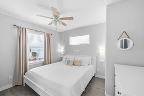 A bedroom with a bed, a ceiling fan, a window with blinds, a bedside table, and a mirror on the wall.