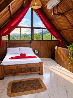 A bedroom with a wooden ceiling, a bed with white linens, red curtains, and a view of greenery outside.