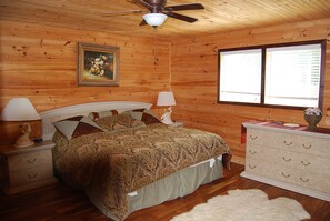 A wooden-paneled bedroom with a bed, two bedside lamps, a dresser, a ceiling fan, and a window with blinds.