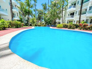 Una piscina amplia y cristalina rodeada de palmeras y un edificio de hotel de varios pisos con balcones.