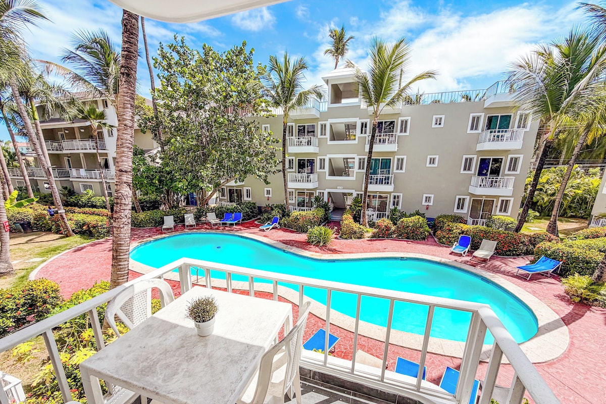 A swimming pool surrounded by lounge chairs and palm trees, with a building in the background.