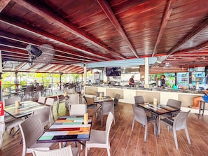 Un restaurante al aire libre con techo de madera, mesas con individuales de colores y una zona de bar con una pantalla de televisión.
