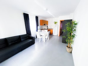 A modern living room with a black sofa, a dining table with chairs, a kitchen area with cabinets, and a potted plant. - Tropical Adventure STUDIOS HOTEL & SPA (Punta Cana)