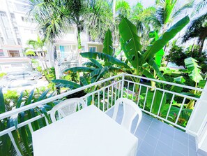 Un balcón con sillas y una mesa blancos, rodeado de exuberante follaje y vistas a una zona residencial.