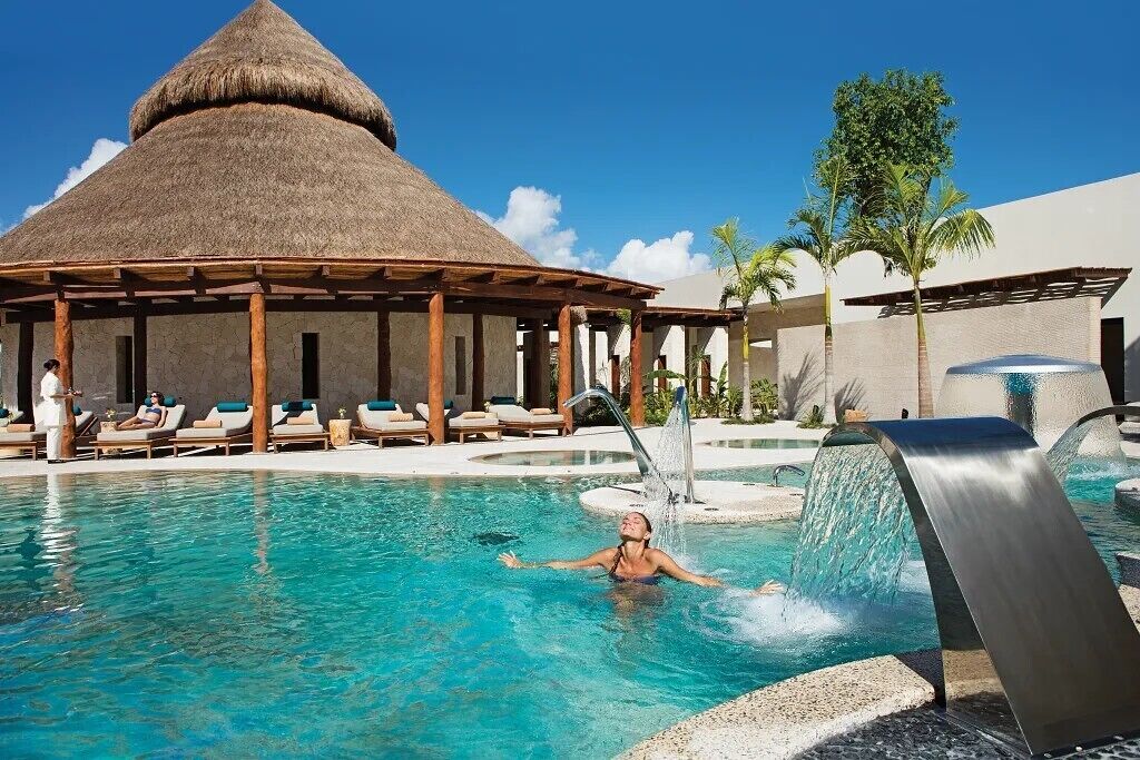 A swimming pool with a water feature, lounge chairs, and a thatched-roof pavilion.