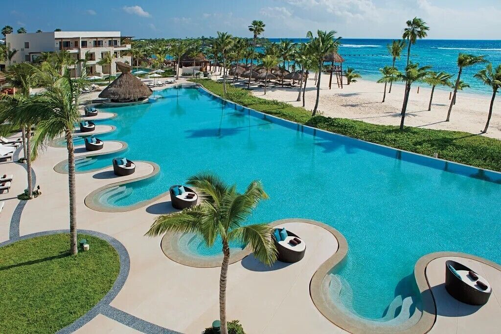 A resort with a large swimming pool, lounge chairs, palm trees, and a beach.