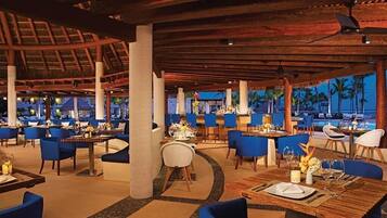 A restaurant with multiple tables and chairs, a thatched roof, and a view of palm trees.