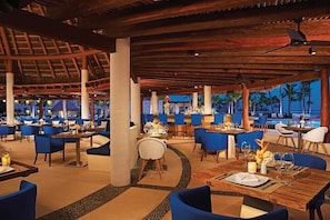 Un restaurante con varias mesas y sillas, techo de paja y vista a palmeras.