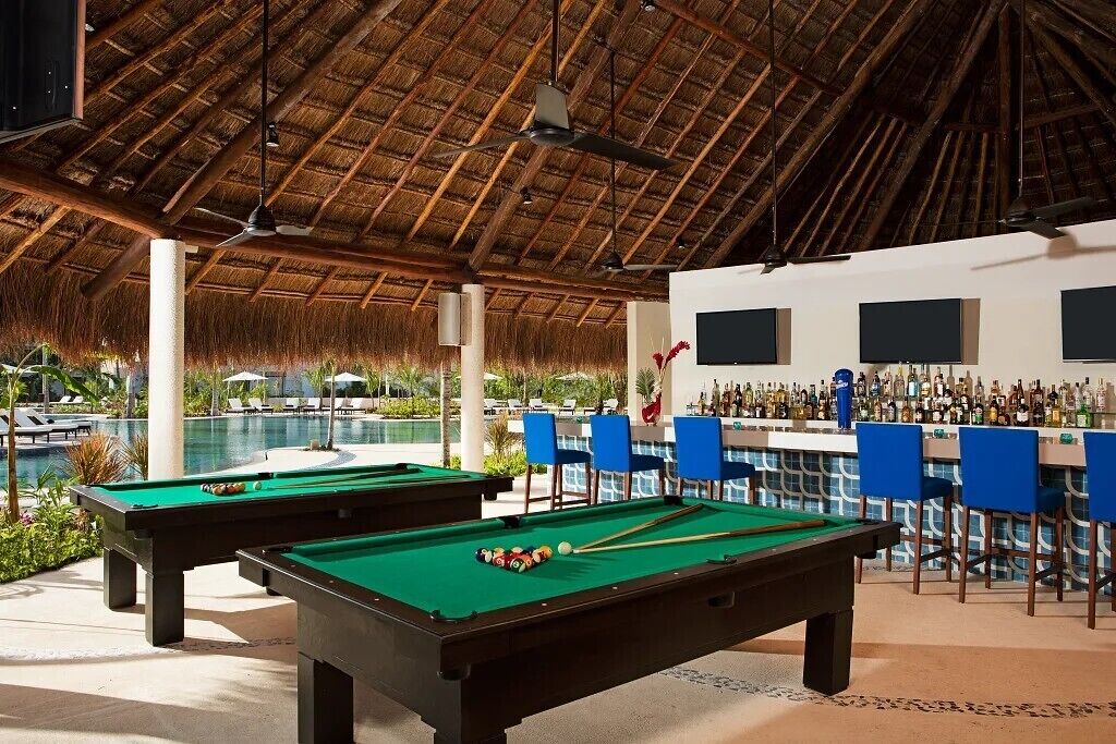 A pool area with two pool tables, a bar with chairs, and a pool.