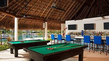 A pool area with two pool tables, a bar with chairs, and a pool.