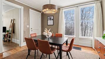 A dining area with a dark wooden table, four chairs, a hanging light fixture, and a view of trees through large windows.