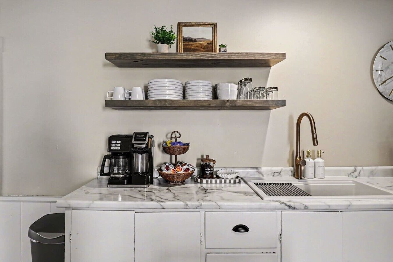 A kitchen with a coffee maker, a sink, and a shelf with plates and cups.