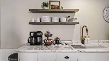 A kitchen with a coffee maker, a sink, and a shelf with plates and cups.