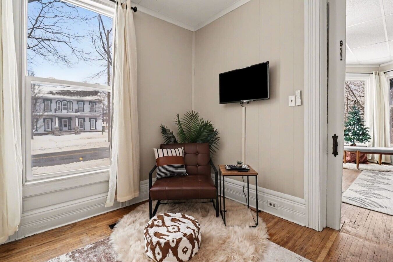 A room with a large window, a brown leather chair, a small table with a plant, and a television on the wall.