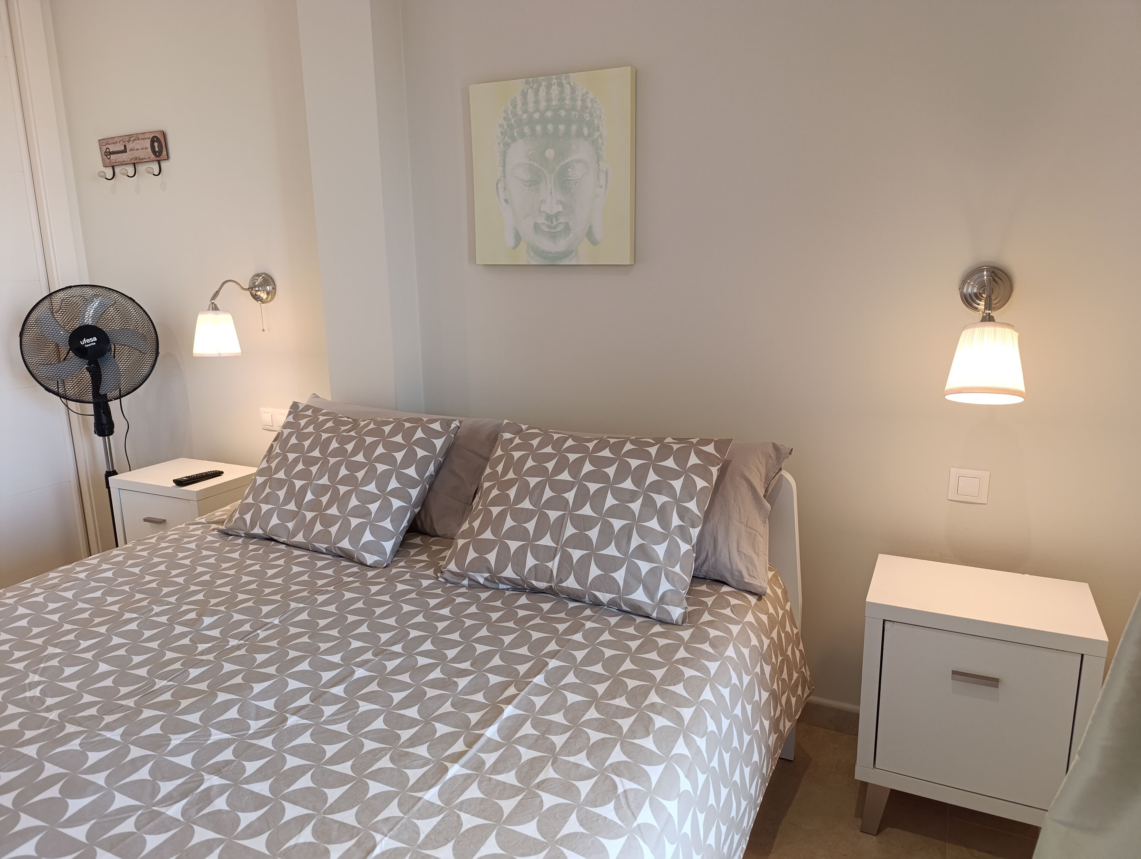 Ein ordentlich bezogenes Bett mit gemusterten Betttextilien, ein Nachttischventilator und Wandlampen.