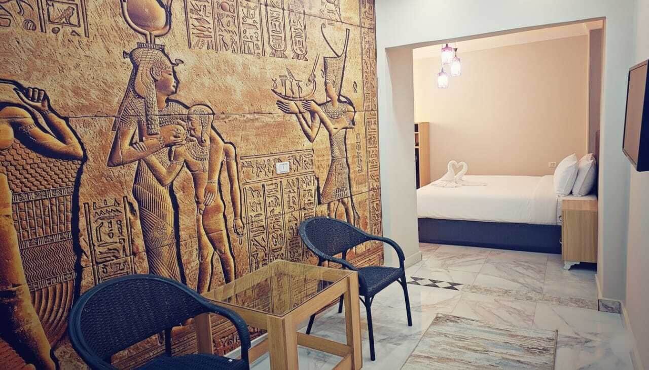 A room with a wall mural of ancient Egyptian hieroglyphs, a bed with white bedding, and a small table with chairs.
