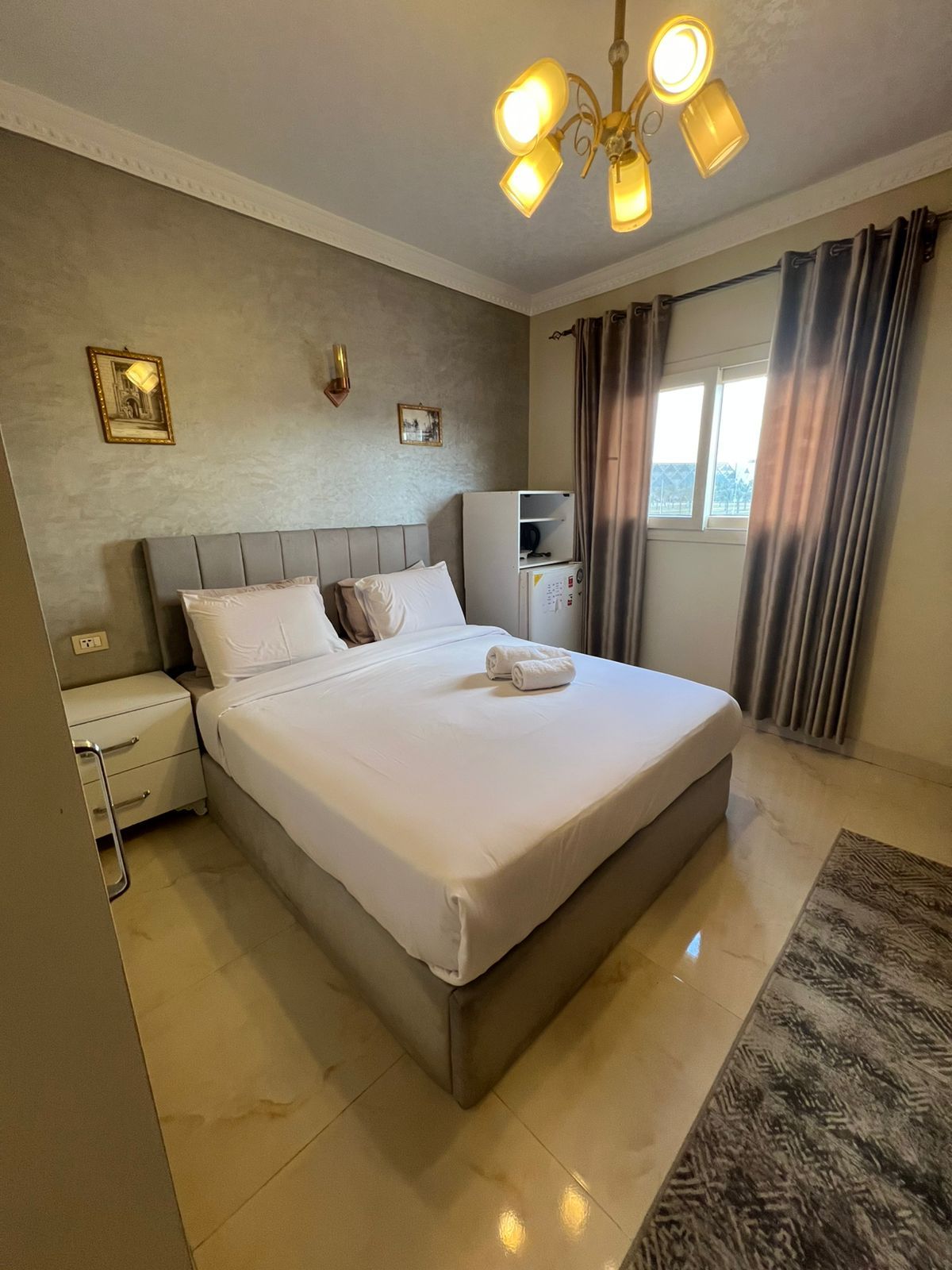 A hotel room with a large bed, a bedside table, a wardrobe, a ceiling light fixture, and a window with curtains.