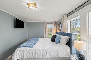 A bedroom with a bed, a TV mounted on the wall, a window with curtains, and a bedside lamp.