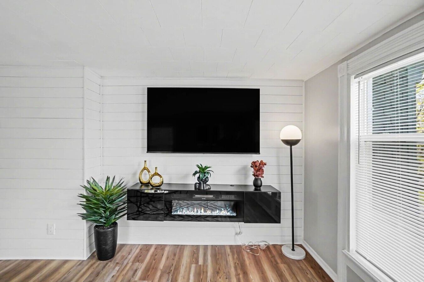 Un salon avec un téléviseur à écran plat fixé au mur, un meuble de rangement noir en dessous et une lampe sur pied à droite.