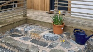 En entré med stenläggning och ett trästaket, en krukväxt med en gul tulpan och en blå vattenkanna.