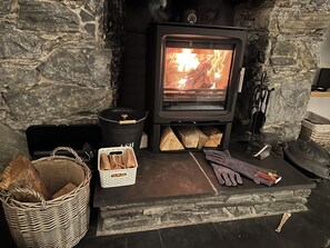 A stone fireplace with a wood-burning stove, logs, a basket, gloves, and a book. - Hawthorn Brae - A Peaceful Highland Retreat (Pitlochry)