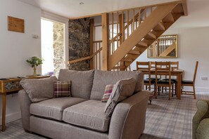A living room with a sofa, a wooden staircase, a dining table with chairs, and a stone wall. - Hawthorn Brae - A Peaceful Highland Retreat (Pitlochry)