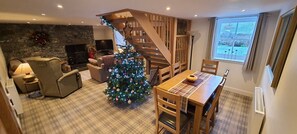 A cozy living room with a fireplace, a wooden staircase, a dining area with a plaid tablecloth, and a Christmas tree. - Hawthorn Brae - A Peaceful Highland Retreat (Pitlochry)
