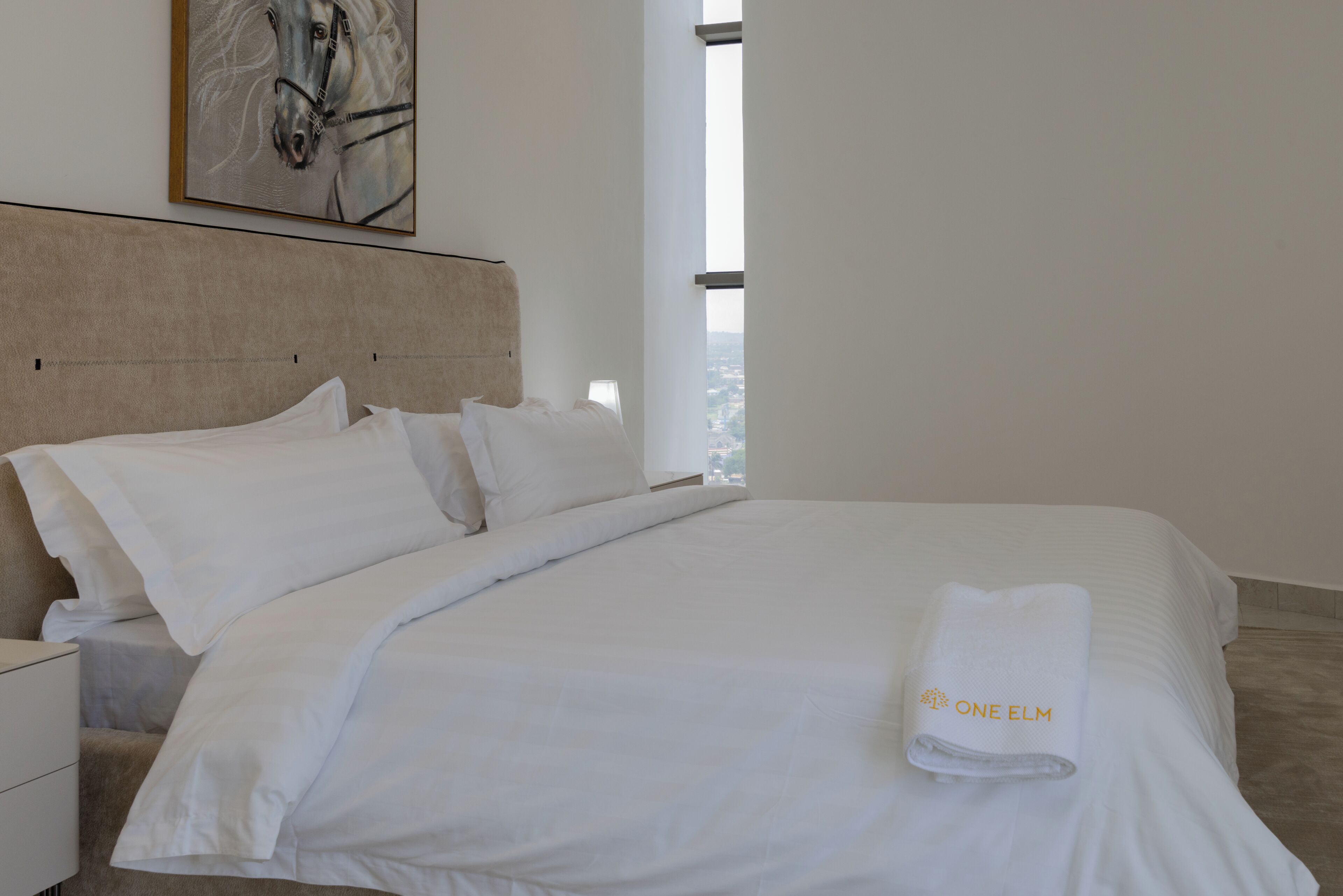 A hotel room with a large bed, white bedding, a headboard, a nightstand, a lamp, and a window with a view.