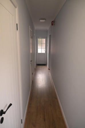 Un pasillo con piso de madera, paredes blancas y una puerta al final.
