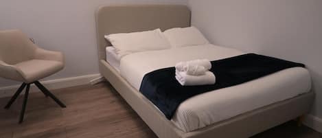 Una cama con ropa blanca y un plaid negro, una silla y piso de madera.