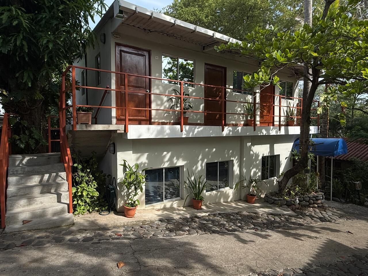 Un edificio de dos pisos con un balcón, plantas en macetas y una escalera que conduce a la entrada.