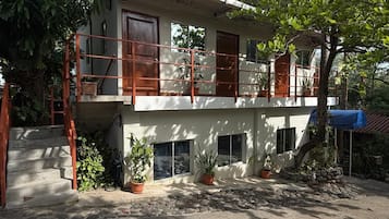 Un edificio de dos pisos con un balcón, plantas en macetas y una escalera que conduce a la entrada.