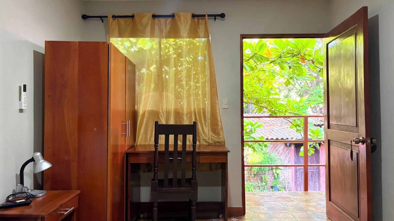 A room with a wooden desk, a chair, a lamp, and a view of a garden through an open door.
