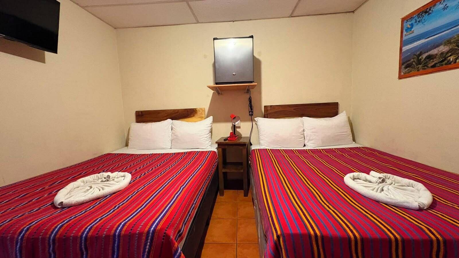 Ein Hotelzimmer mit zwei Einzelbetten, jeweils mit einer rotgestreiften Bettdecke und weißen Kissen. Zwischen den Betten steht ein kleiner Tisch mit einer Lampe, und ein Fernseher ist an der Wand über dem Tisch angebracht.