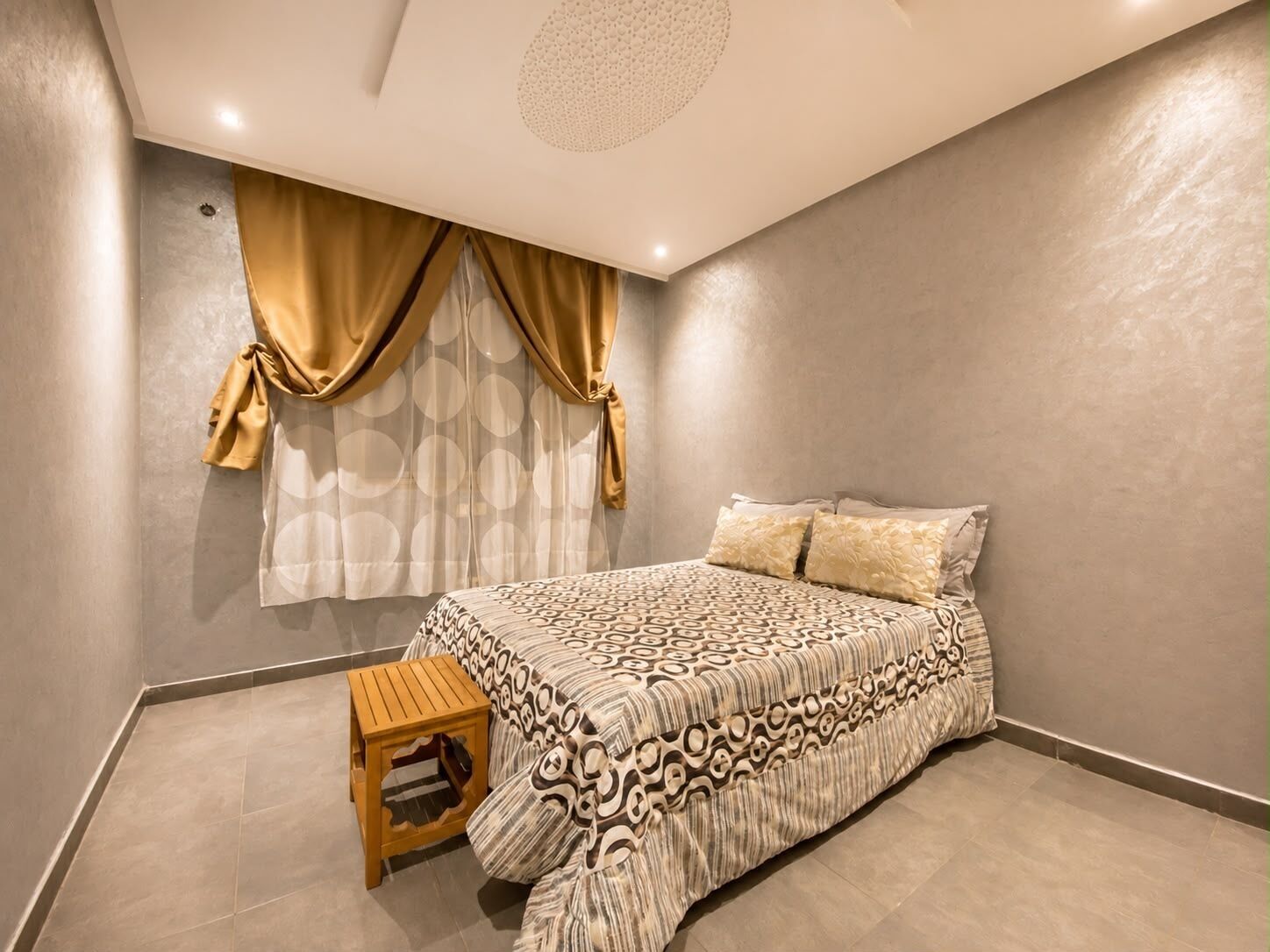 Une chambre à coucher avec un lit, des rideaux, un petit tabouret en bois et un luminaire fixé au plafond.