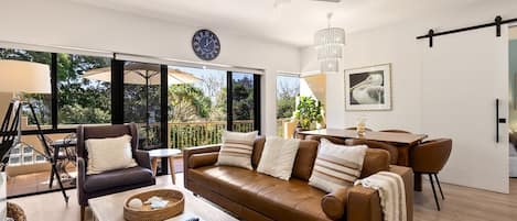 A modern living room with a brown leather sofa, wooden coffee table, and a dining area with a chandelier.