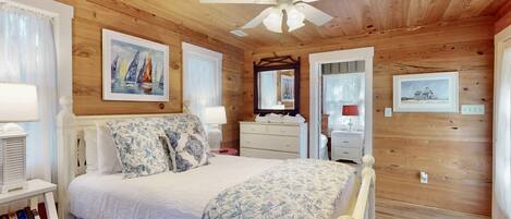 A bedroom with a wooden ceiling, a bed with floral bedding, a nightstand with a lamp, a bookshelf, and a ceiling fan.