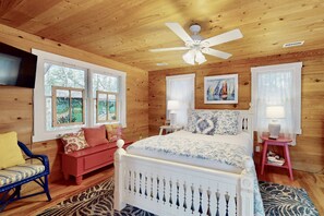 Quarto de cabana de madeira com cama, sofá vermelho, cadeira azul e ventilador de teto.