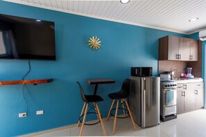 A modern kitchen with a blue wall, a flat-screen TV mounted above a wooden bar counter, a refrigerator, and a microwave. - Aruba Vibe Apartments 9 | Charming Studio Near the Beaches and Downtown (Oranjestad)