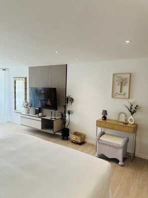 A modern bedroom with a large bed, a TV mounted on a wall, a small table with decorative items, and a wicker bench. - Bohemian studio just a stone's throw from the beach (Mont vernon)