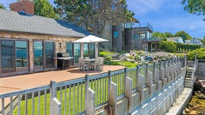 A deck with outdoor furniture, a grill, and a stone building with large windows. - Stay at Windward Point (Gloucester)